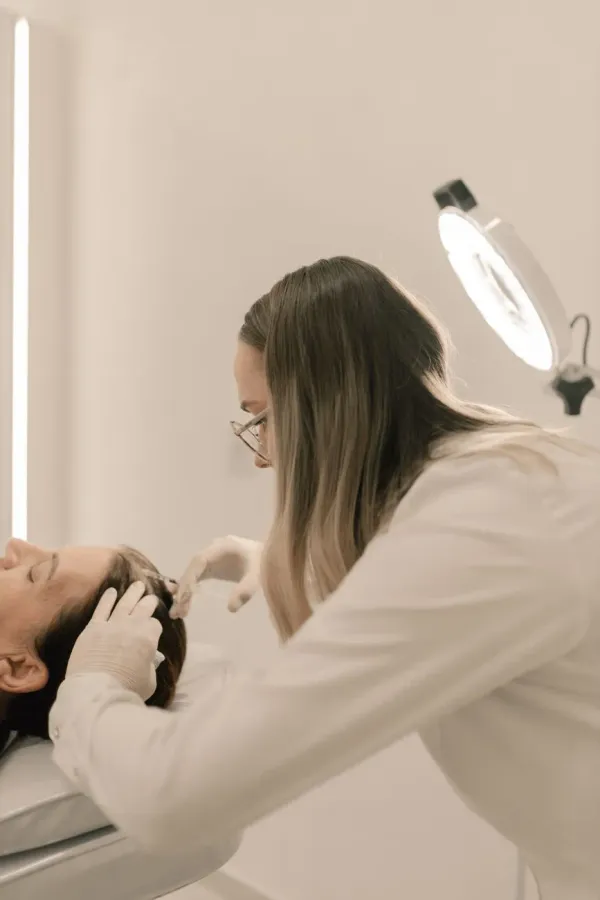 A practitioner with glasses leans in to look closely at a client's skin in a softly lit clinic room