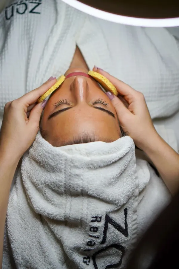 A client lies back with a towel wrapped around her hair while a practitioner cleanses her face with sponges